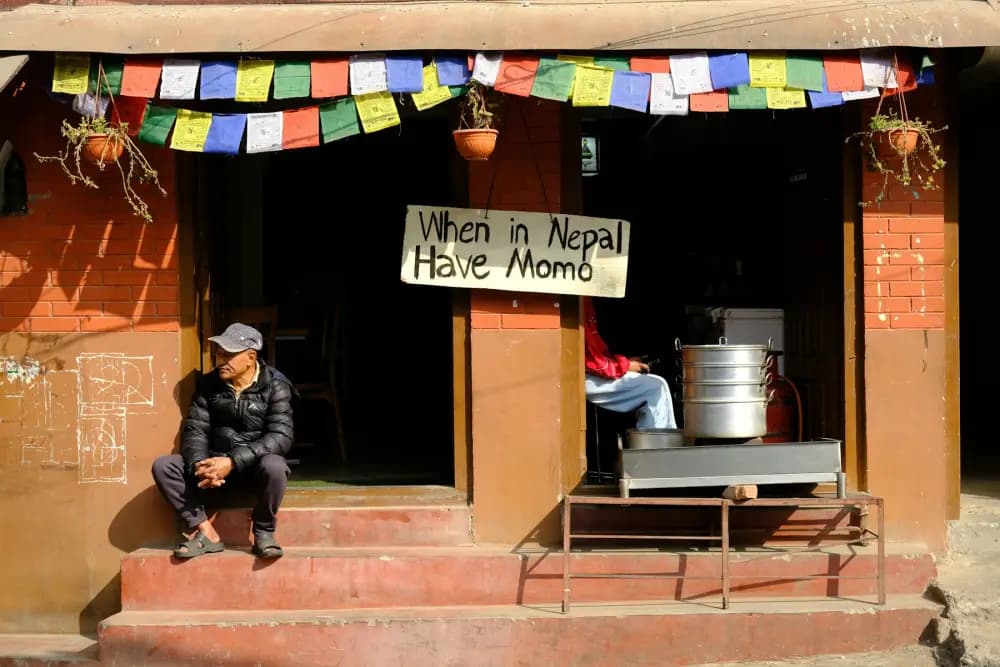 A man sitting on the steps of a small momo restaurant decorated with prayer flags